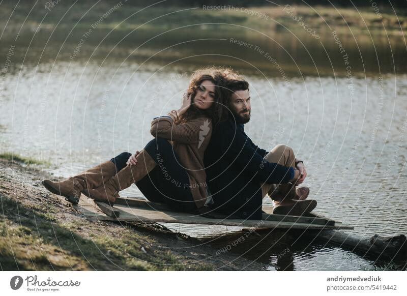 two caucasian lovers sitting on the pier by the lake. Young couple is hugging on autumn day outdoors. A bearded man and curly woman in love. Valentine's Day. Concept of love.