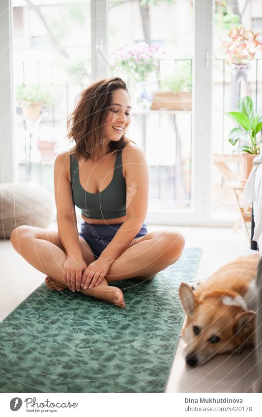 young woman with corgi Pembroke dog doing yoga on green mat at home. Practicing sport, wellness and healthcare. Indoor yoga online class. Mindfullnesss and meditation, active lifestyle.