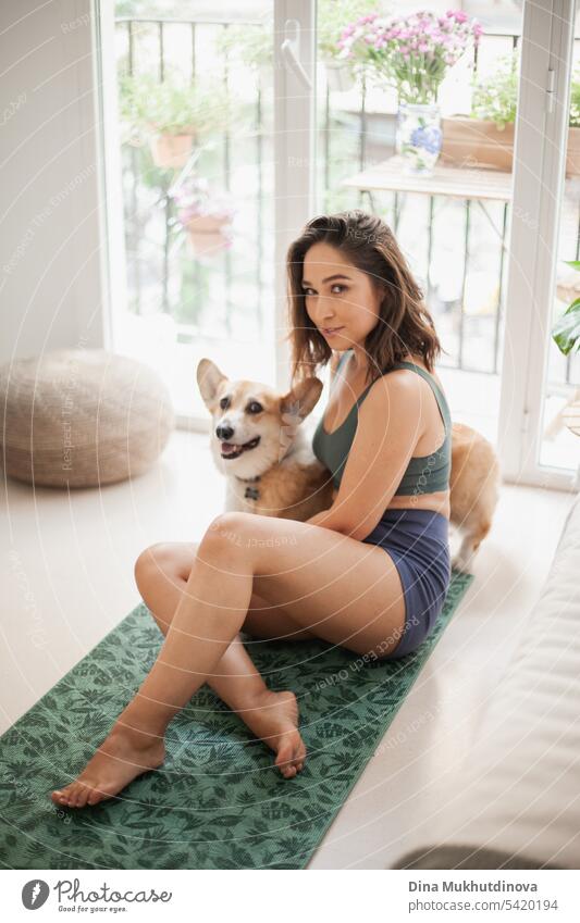 young woman with corgi Pembroke dog doing yoga on green mat at home. Practicing sport, wellness and healthcare. Indoor yoga online class. Mindfullnesss and meditation, active lifestyle.