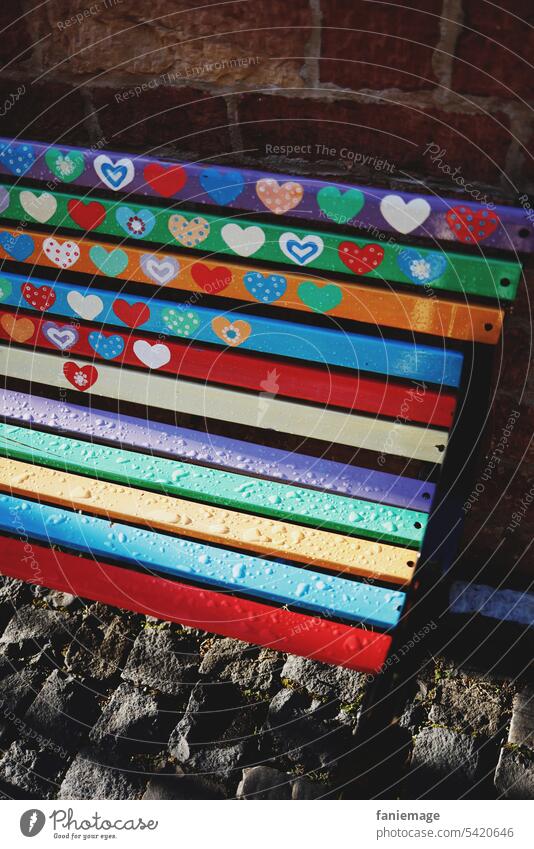 mit Herzen versehene Bank in Pride- Farben Parkbank Regenbogenfarben Regentropfen Herzchen Fußgängerzone LGBTQ LGBTQA+ Symbol bunt gestreift Kopfsteinpflaster
