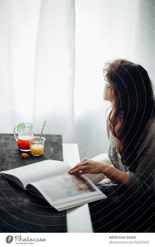 Young woman reading book at table with juice drink rest home relax pensive young female lifestyle literature casual morning window hobby fresh enjoy peaceful