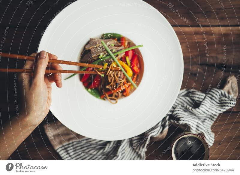 Person eating Asian noodles with chopsticks food dish asian hand meat vegetable meal delicious wooden white plate dinner tasty lunch cuisine healthy cook