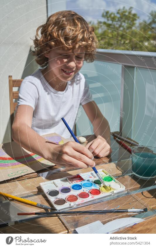 Cheerful boy painting on balcony watercolor table smile container brush art creative kid child draw childhood tool sit happy lifestyle home apartment sunny