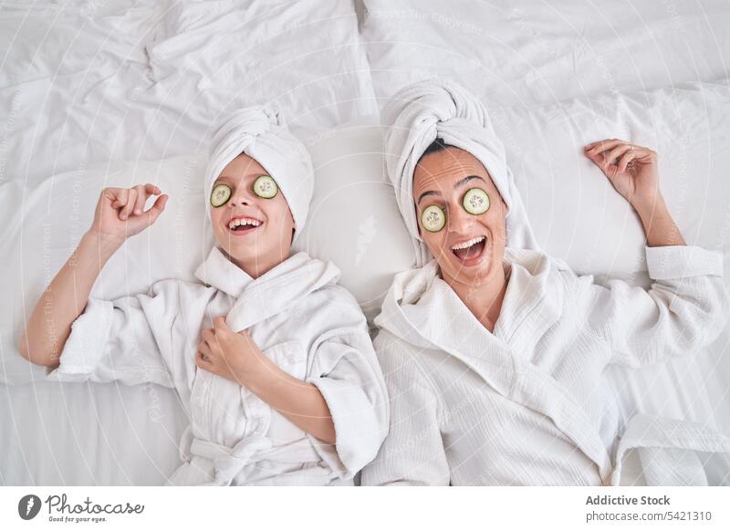 Cheerful woman with kid enjoying spa procedure at home beauty skin care cucumber mask mother together having fun laugh rest cozy positive facial natural smile