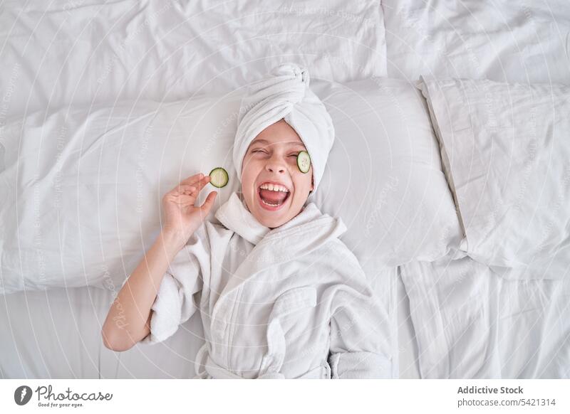 Cheerful kid with slices of cucumber on yes lying on bed mask beauty skin care fun positive laugh enjoy spa facial joke humor playful natural child happy