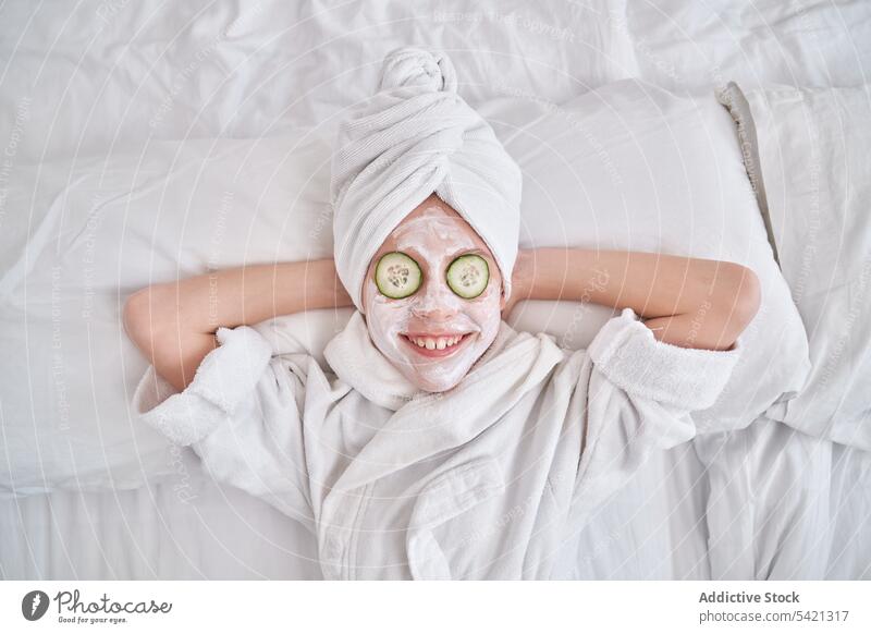 Playful kid with facial mask lying on bed having fun cucumber beauty skin care grimace playful show tongue enjoy spa joke humor natural child happy make face