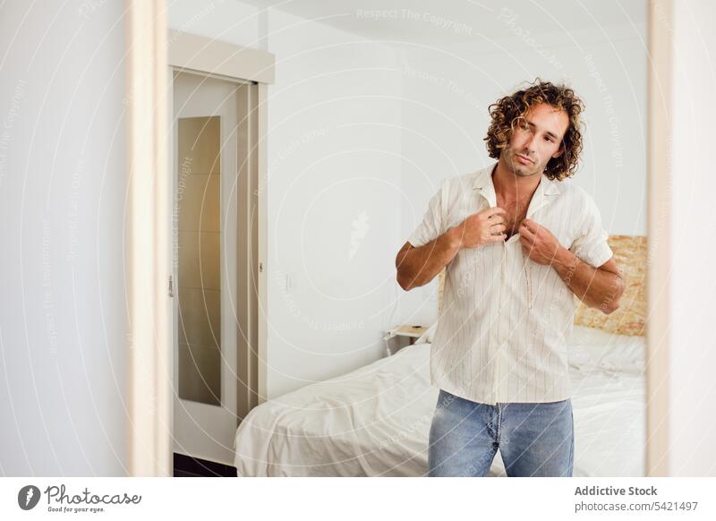 Handsome man buttoning shirt in room put on dress up reflection mirror style handsome outfit male home modern prepare apartment flat stand guy curly hair