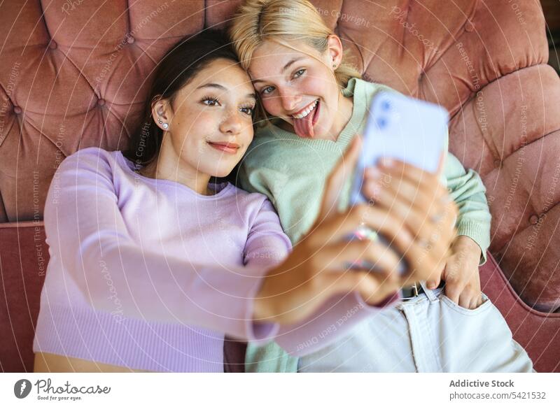 Content teenage sisters taking selfie on smartphone on couch self portrait girl mobile tender young sofa using lying device smile delicate gentle delight