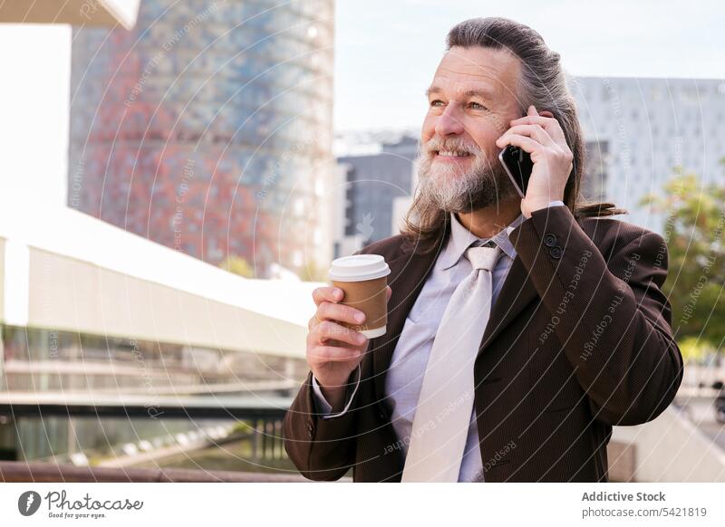 Cheerful bearded businessman with coffee talking on smartphone on street urban smile takeaway modern mature middle age gray hair style elegant cup drink