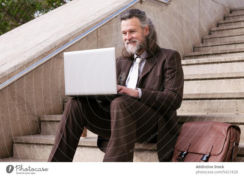 Smiling mature man in suit using laptop on stairs businessman online remote style happy gadget internet communicate entrepreneur positive smile middle age aged