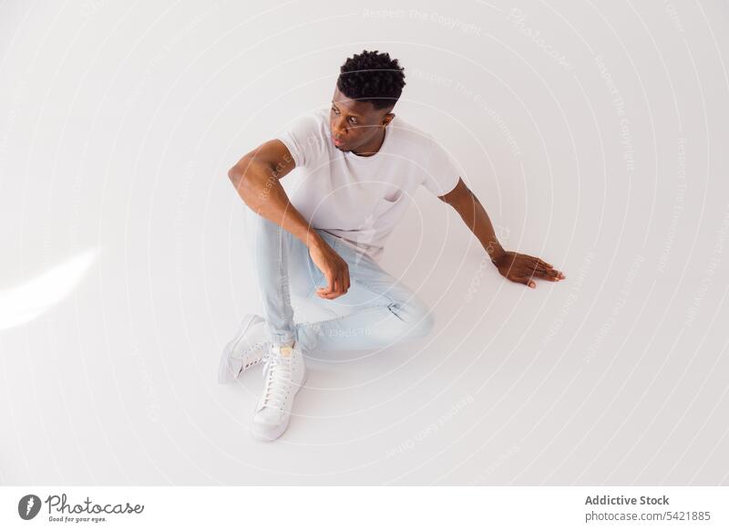 Black man in white outfit sitting in studio style fashion casual summer trendy look modern male model black african american ethnic afro t shirt jeans sneakers
