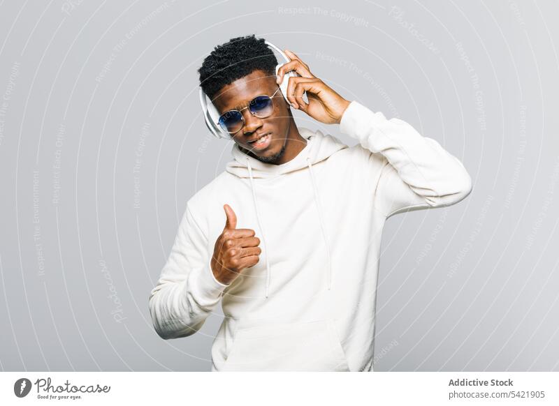 Cheerful black man in trendy outfit listening to music headphones thumb up style hipster white sunglasses hoodie fashion young african american gesture approve