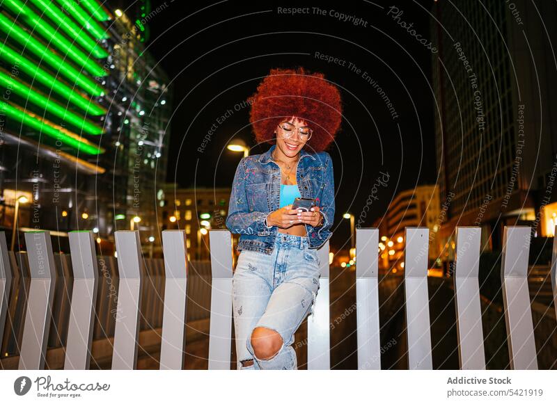 Stylish woman browsing smartphone on dark street with buildings online afro evening style fencing fashion urban rendy apparel surfing cellphone female gadget