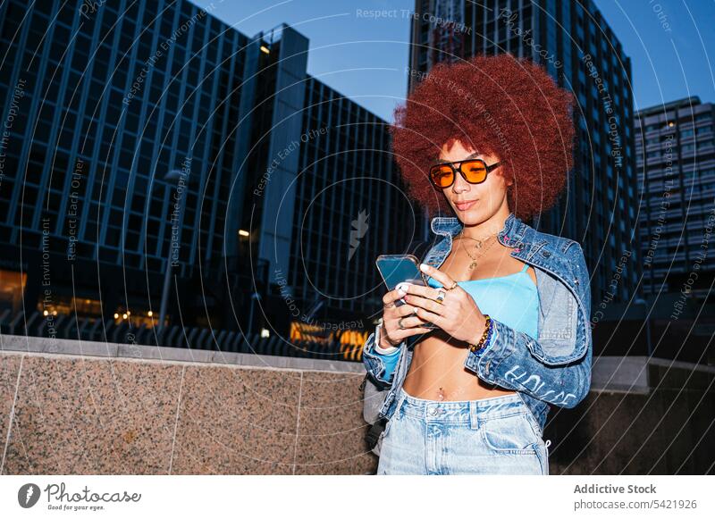 Stylish woman browsing smartphone on street online afro evening building urban fashion house style trendy apparel surfing text message cellphone female barrier
