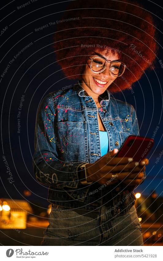 Cheerful woman browsing smartphone on dark street online afro evening building style fashion urban house trendy apparel surfing cellphone female gadget device