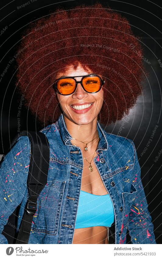 Happy woman in stylish wear on dark street denim afro style fashion apparel street style backpack evening twilight female hairstyle sunglasses eyewear design