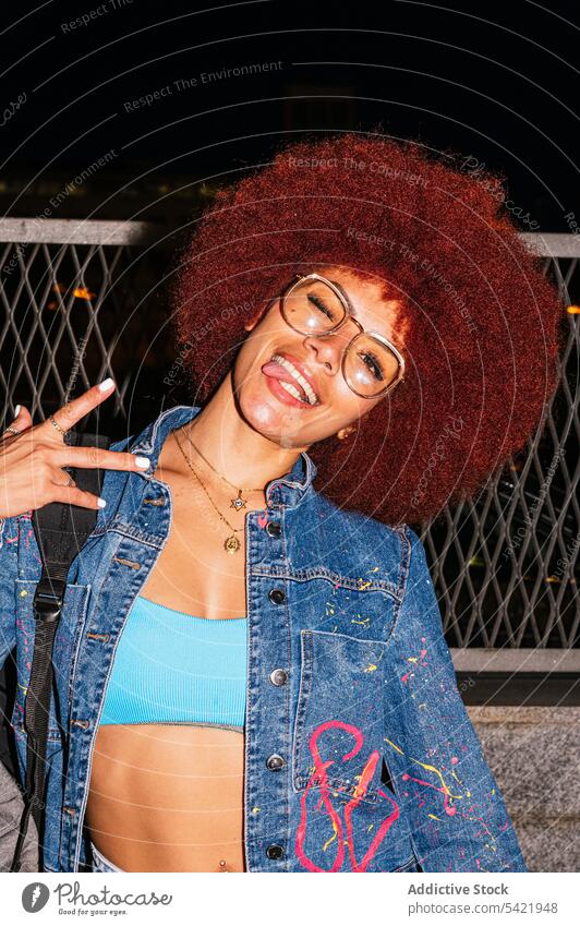 Stylish woman showing peace gesture on dark street afro style fashion apparel outfit evening v sign carefree twilight appearance female hairstyle tongue out