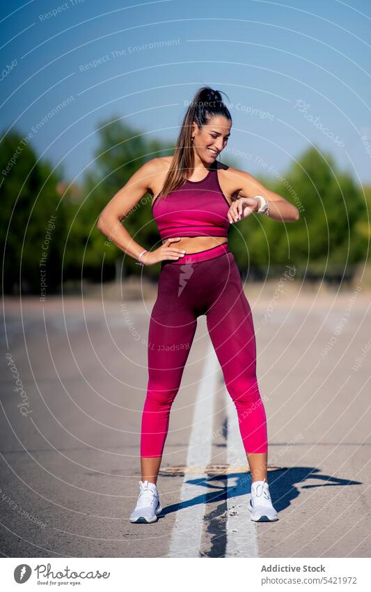 Cheerful female athlete on asphalt looking at fitness tracker woman bracelet smart watch sportswear training sporty slim activewear lifestyle summer workout