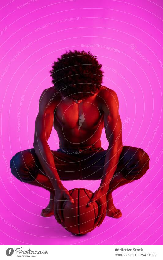 Black shirtless man sitting with basketball naked torso muscular sportsman appearance player neon squat male african american black curly hair young confident