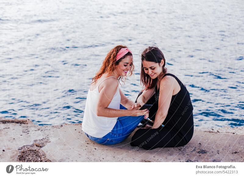 two women friends sitting by the beach using mobile phone and smiling. Technology and lifestyle concept casual stylish cheerful positive sunlight looking