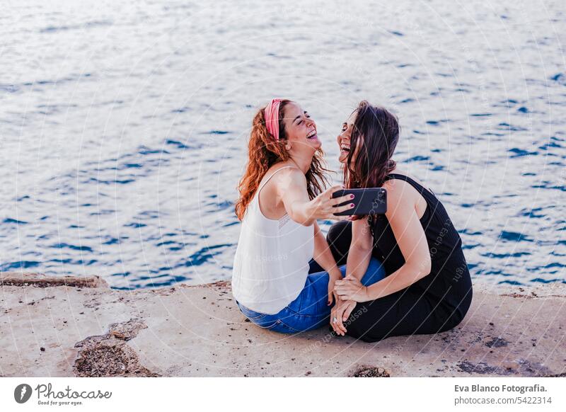 two women friends sitting by the beach using mobile phone, taking a selfie and smiling. Technology and lifestyle concept casual stylish cheerful positive