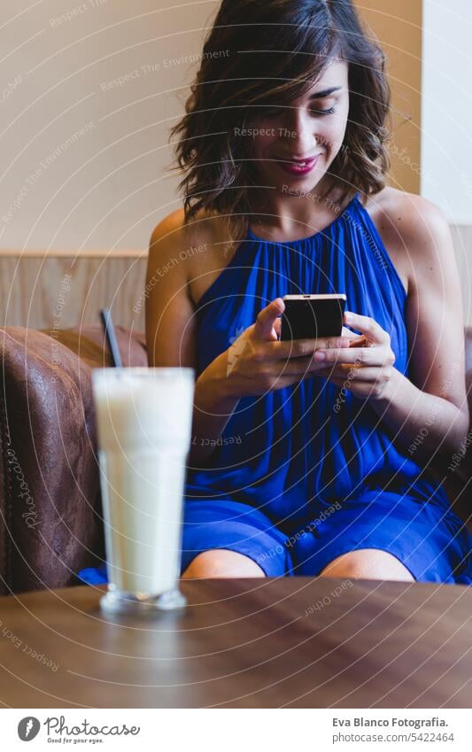 young beautiful woman enjoying a smoothie drink in a cafe and using mobile phone. Casual blue dress. Modern life indoors. selective focus person attractive
