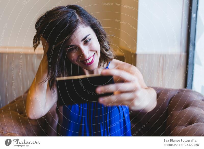 young beautiful woman taking a selfie with mobile phone and smiling. wearing a casual blue dress. Indoors, technology and lifestyle portrait person camera