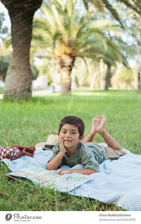 diverse kid reading a book in the park on the blue blanket on green grass, smiling and looking to the camera. Five or six years old boy in the park in summer.