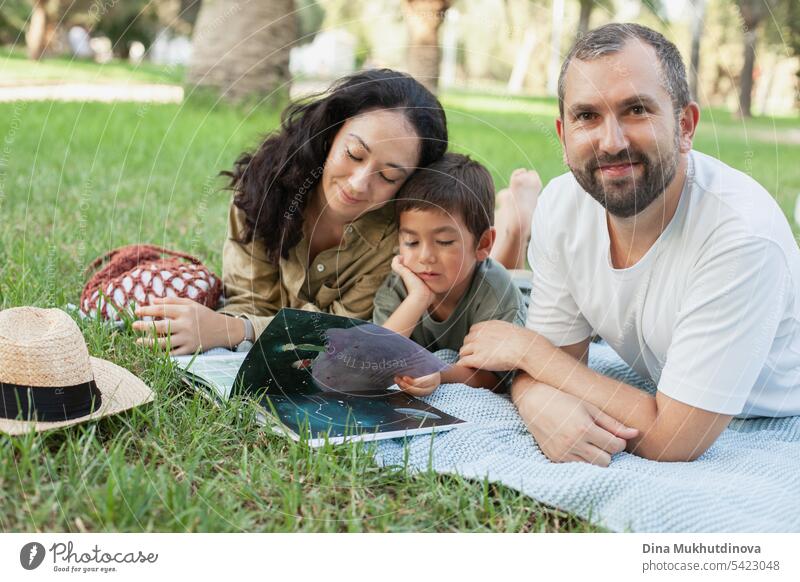 happy family smiling with a son reading a book together in park outdoors in summer mom dad father mother parenthood parents fatherhood motherhood love child