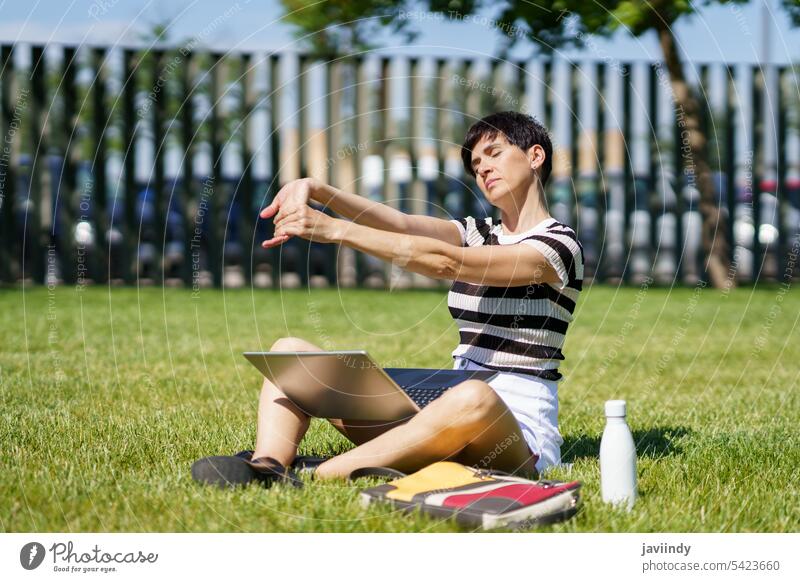 Female student stretching on green lawn in park and laptop with folder woman freelance pc eyes closed bottle netbook grass sit exercise drink online project