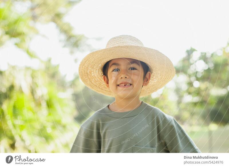 diverse kid standing in the park, wearing straw hat, smiling and looking to the camera. Five or six years old boy in the park in summer with palm trees as background.