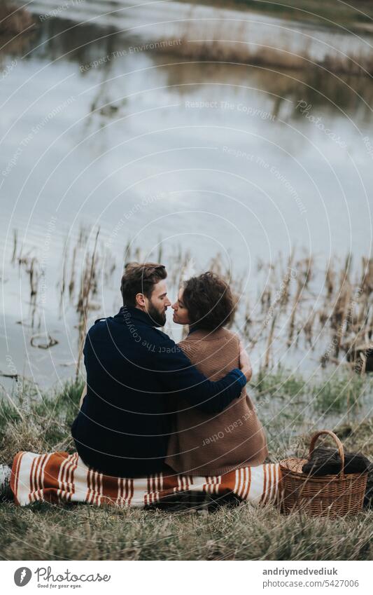two caucasian lovers sitting on blanket by the lake. Young couple is hugging on autumn day outdoors. A bearded man and curly woman in love. Valentine's Day. Concept of love and family. back view