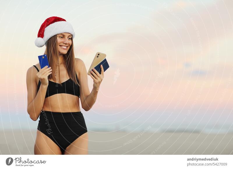 Happy young woman in Santa hat and swimsuit with smartphone, credit card and passport is booking online ticket to the travel Christmas and New Year holiday vacation on beach. Discount trips concept.