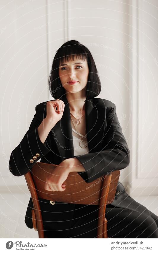 attractive business woman sits on wooden chair and looking in camera. brunette lady with short hair wears black office suit and white blouse, high-heels on white wall background. copy space.