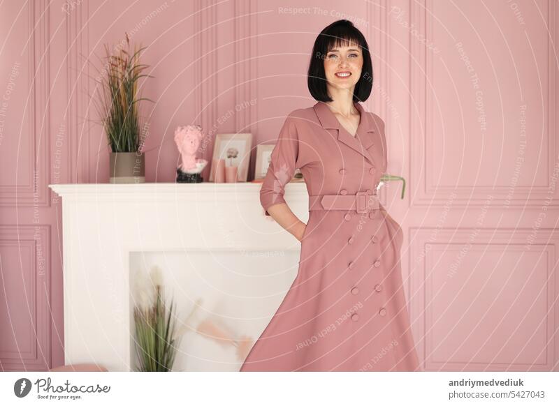 Portrait of attractive smiling brunette with short hair woman folded arms staying alone at stylish interior indoors. Female in soft pink dress is looking in camera