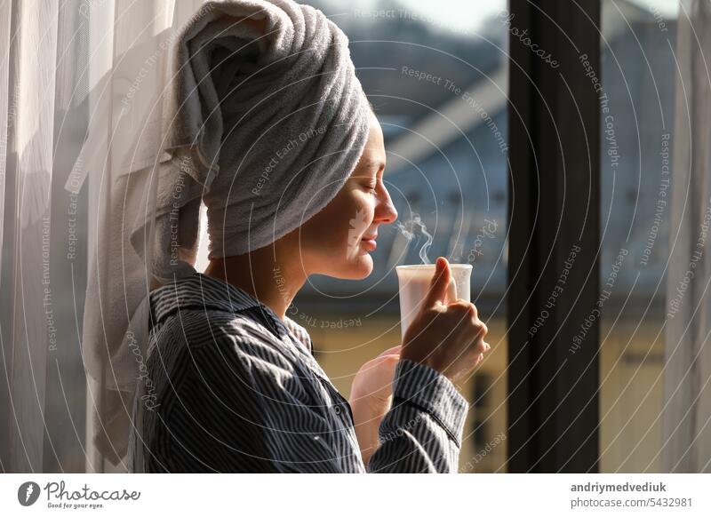 Beautiful young woman with bath towel on head holds a white cup of hot coffee or tea at home by the window. Smiling girl after shower is enjoying aroma drinks and have breakfast in the morning