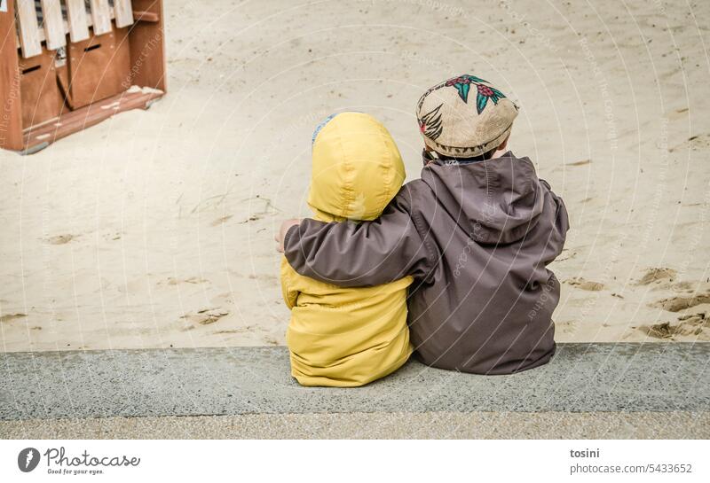 Two children sit on the beach and watch out for each other Brothers Connectedness Beach Sand Sandy beach Rain jacket take care of each other Friendship