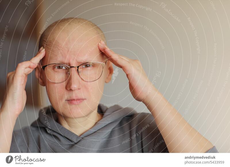 Hairless woman in glasses touching head with hands, suffering from headache after chemotherapy. Unhappy bald cancer sick female patient having migraine at home. Oncology concept. Healthcare, medicine