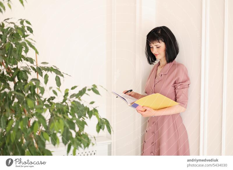 Young brunette woman in stylish soft pink dress is looking in yellow folder of paperwork with mock up area and standing in office with houseplants. Education, business, lifestyle concept