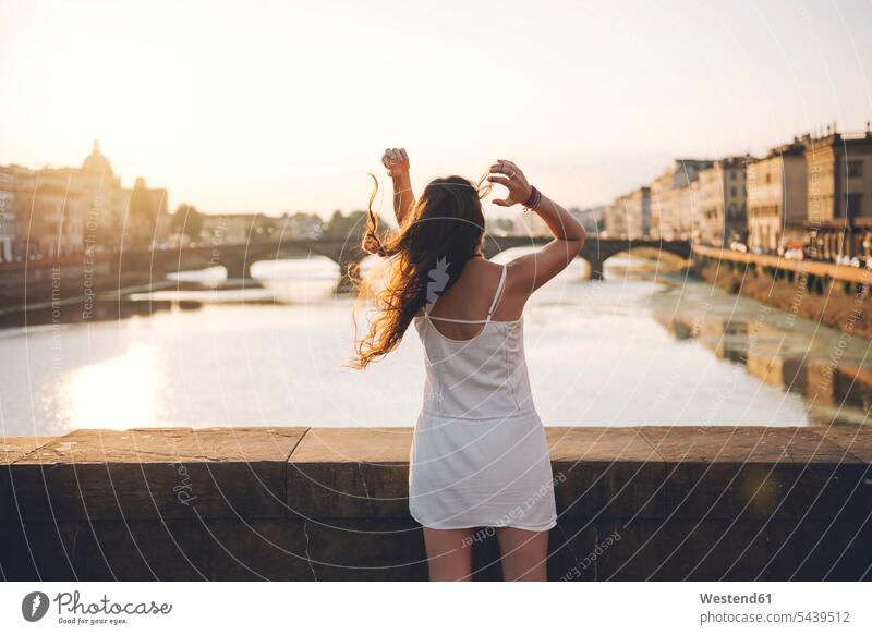 Italy, Florence, back view of woman wearing white summer dress standing on a bridge at sunset Backlit back lighting backlight happiness happy long-haired