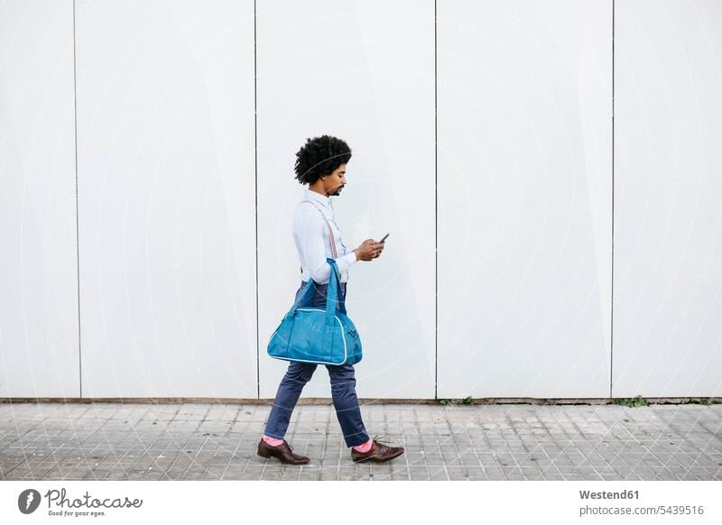 Man with bag walking in front of a white wall while looking at cell phone man men males bags walls Smartphone iPhone Smartphones eyeing going Adults grown-ups
