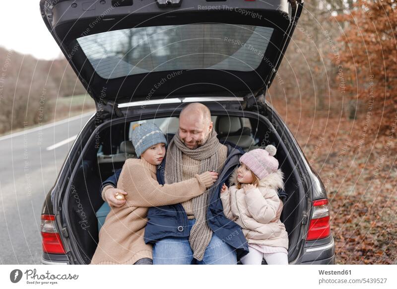 Smiling father with two children sitting in car trunk at the roadside automobile Auto cars motorcars Automobiles pa fathers daddy dads papa Road Side boot