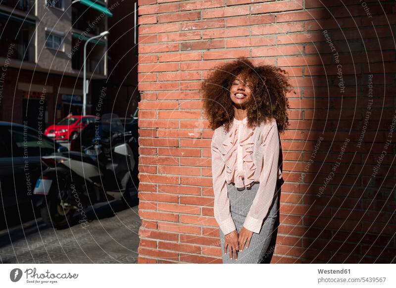 Portrait of smiling young woman with afro hairdo leaning against brick wall in the city portrait portraits smile Afro town cities towns females women beautiful