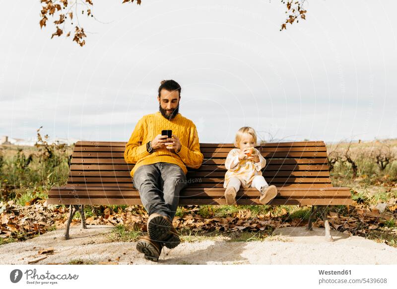 Father and daughter sitting on a bench in the park in autumn, father using smartphone, daughter eating an apple morning in the morning use Smartphone iPhone