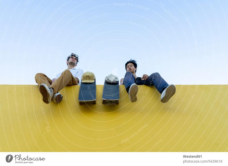 Two young men with skateboards sitting on a high yellow wall friends mate Skate Board resting walls man males Seated friendship Adults grown-ups grownups adult
