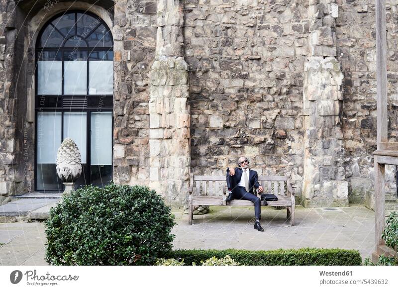 UK, London, senior businessman sitting on bench in a courtyard relaxing while liestening music with headphones relaxation hearing patio Seated benches headset
