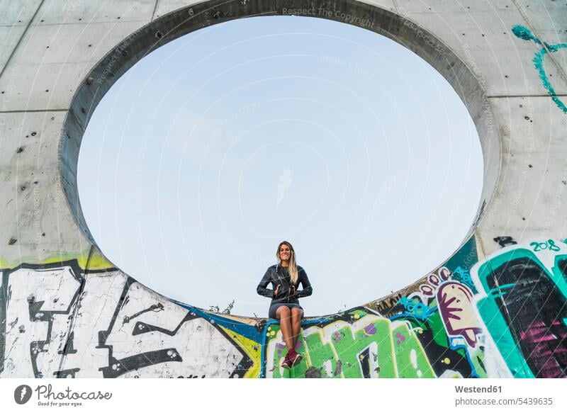 Smiling young woman sitting in a large hole in a concrete wall concrete walls Seated holes big females women size Adults grown-ups grownups adult people persons