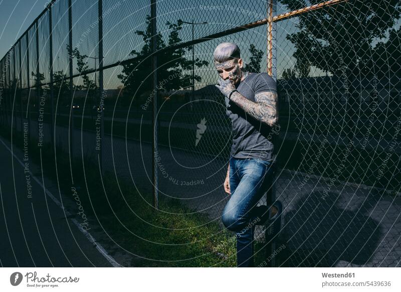 Tattooed young man smoking a cigarette at wire mesh fence men males cigarettes tattooed Wire Mesh Fence Chainlink Fence Chain Link Fence Chain-link Fence