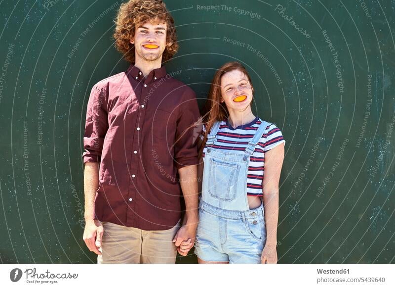 Young couple standing in front of a green wall with orange slices in their mouth Slice Slices Orange Oranges walls twosomes partnership couples colour colours