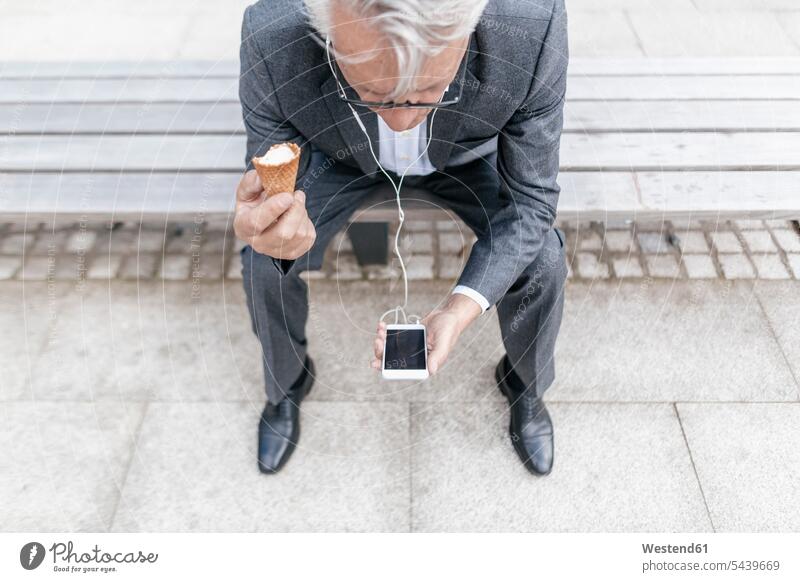 Senior businessman with ice cream cone sitting on bench looking at cell phone Businessman Business man Businessmen Business men business people businesspeople